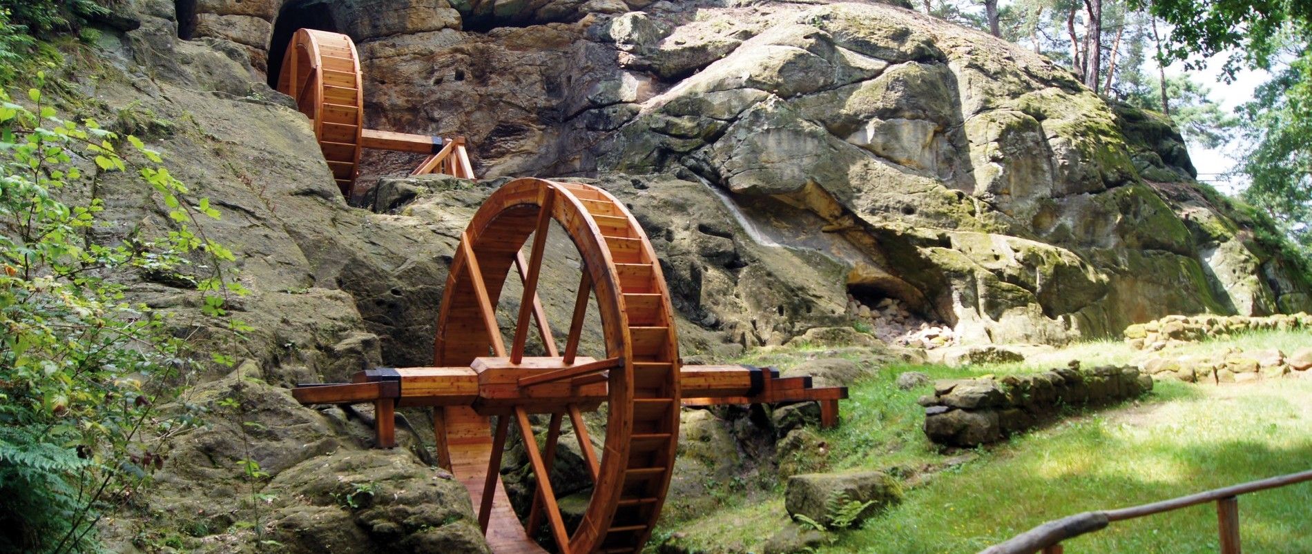 Zwei hölzerne Wasserräder an einem Felsen in einer grünen Umgebung.