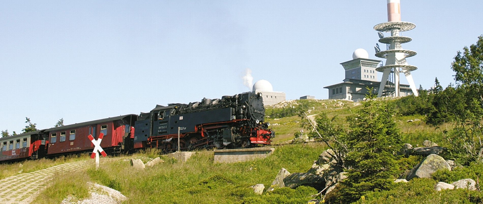 Eine historische Dampflokomotive zieht einen Zug am Brocken, mit Aussichtsturm und Gebäuden im Hintergrund.