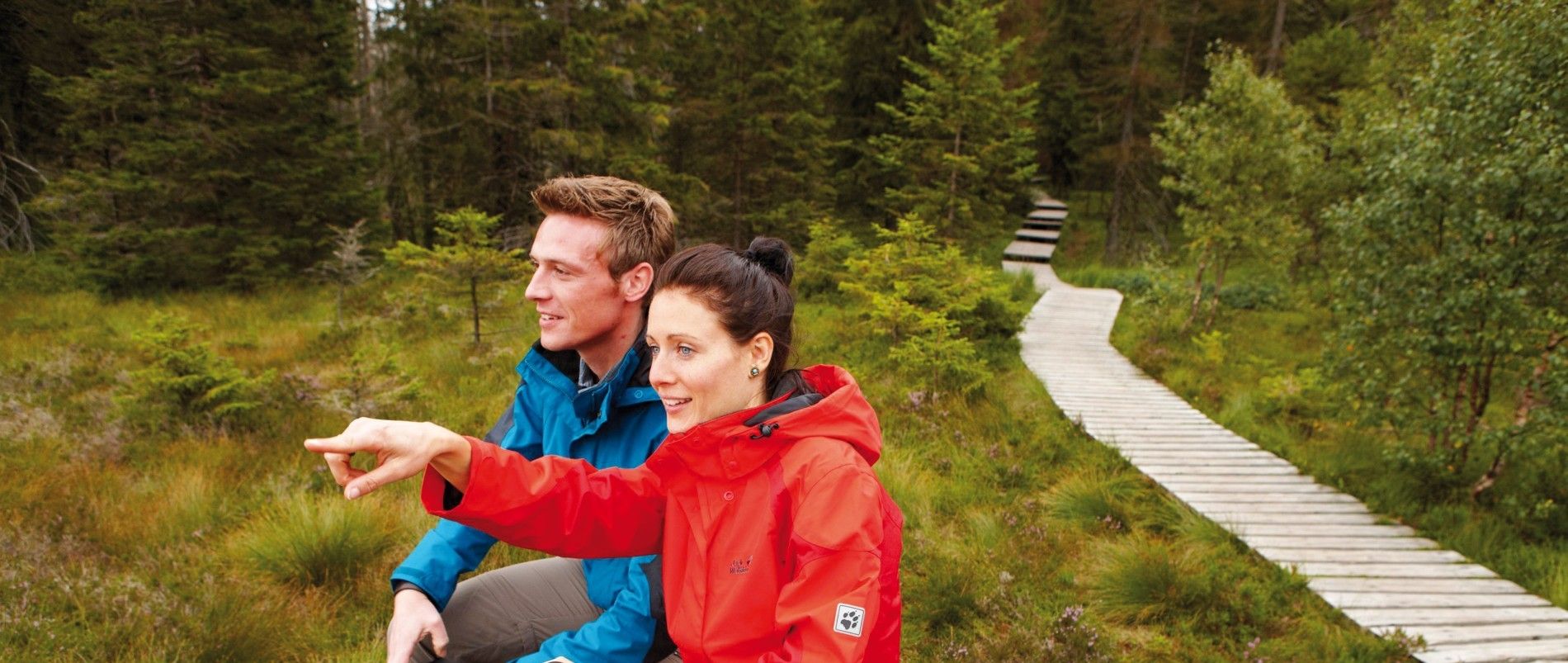 Zwei Personen in Outdoor-Kleidung sitzen auf einem Holzsteg in einem bewaldeten Gebiet und schauen in die Ferne, während eine der Personen mit dem Finger zeigt.