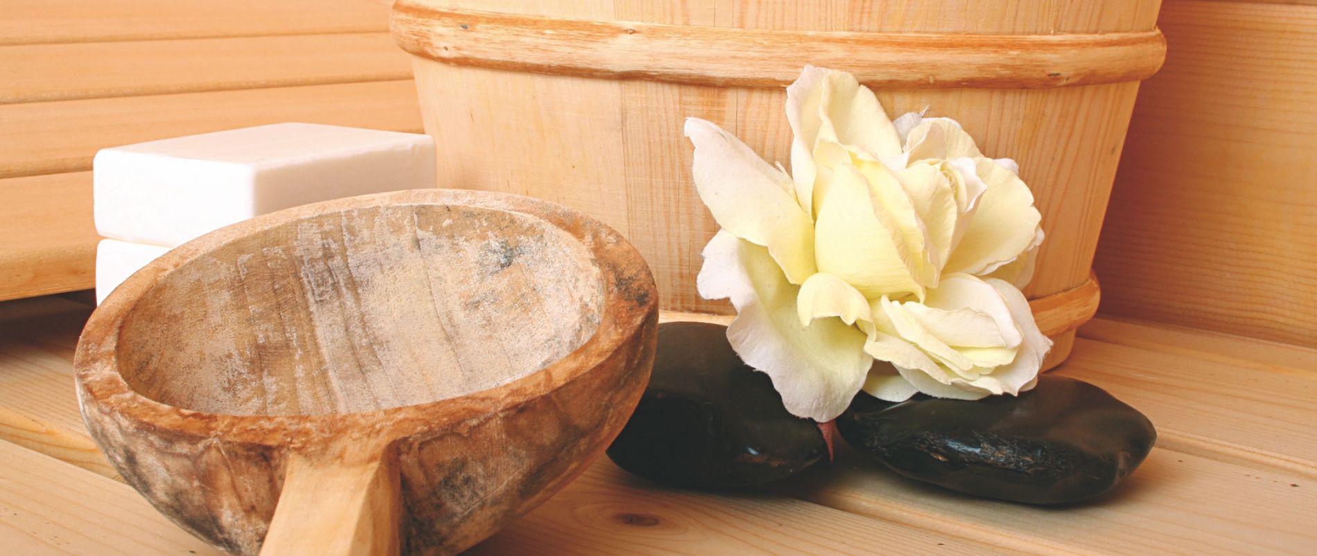 Sauna-Accessoires mit Holzschöpfkelle, Holzfass, helle Blume und Steinen auf Holztisch