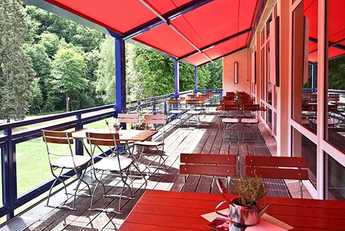 Außenterrasse eines Restaurants mit roten Sonnenschirmen, Holztischen und Stühlen, umgeben von grüner Natur.