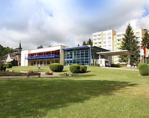 Gebäudekomplex mit modernem Design, Rasenfläche im Vordergrund und Bäumen im Hintergrund.