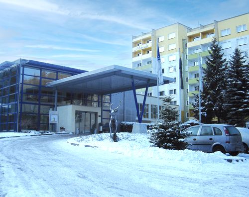 Verschneites modernes Gebäude mit blauem Glas und angrenzenden Wohnkomplex, umgeben von schneebedeckten Bäumen und geparkten Autos.