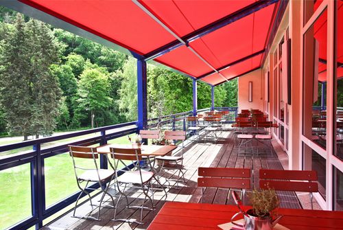 Holzterrasse mit roten Markisen, Bistrotischen und Stühlen, mit einem Blick auf grüne Bäume.