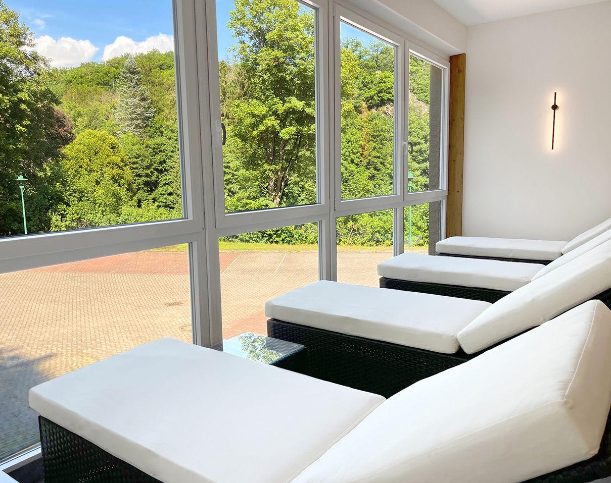 Ein helles Zimmer mit großen Fenstern, das auf eine grüne Landschaft blickt. Im Raum stehen mehrere Liegestühle mit weißen Polstern.