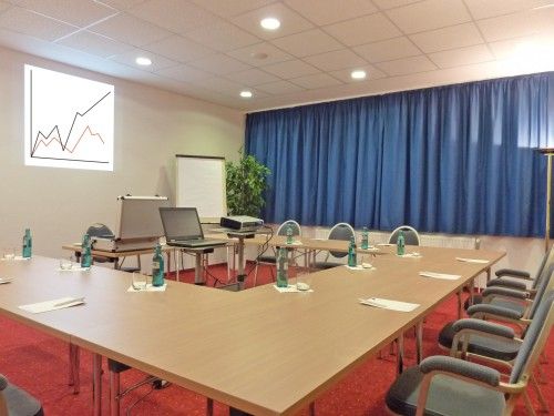 Konferenzraum mit einem Tisch in U-Form, ausgestattet mit Wasserflaschen, Gläsern und Schreibmaterial, sowie einem Beamer und einem Laptop. An der Wand ein Diagramm.