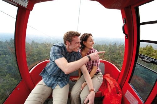 Zwei Personen sitzen lächelnd in einer roten Seilbahnkabine und zeigen auf die Landschaft.