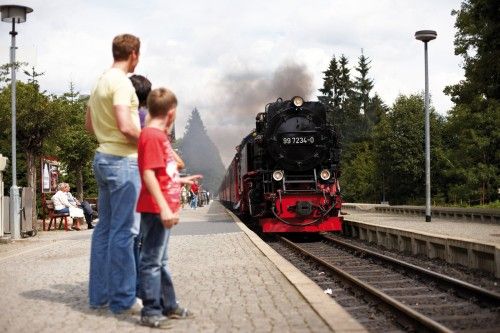 Eine Dampflokomotive fährt in einen Bahnhof ein, während mehrere Personen am Bahnsteig stehen und zuschauen.