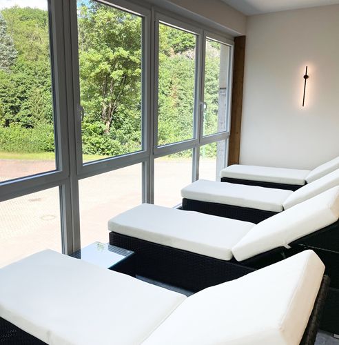 Innenraum mit mehreren weißen Liegen vor großen Fenstern und Blick ins Grüne.