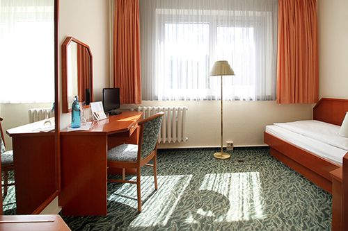 Ein einfach eingerichtetes Hotelzimmer mit einem Einzelbett, Schreibtisch und Stuhl, unter einem Fenster mit orangefarbenen Vorhängen.