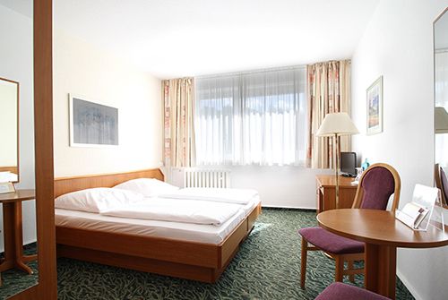 Helles Hotelzimmer mit Doppelbett, Tisch, Stuhl, Fernseher und großen Fenstern.