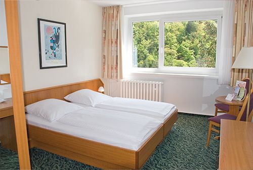 Helles Hotelzimmer mit einem Doppelbett, Kunst an der Wand und einem großen Fenster mit Blick auf grüne Bäume.
