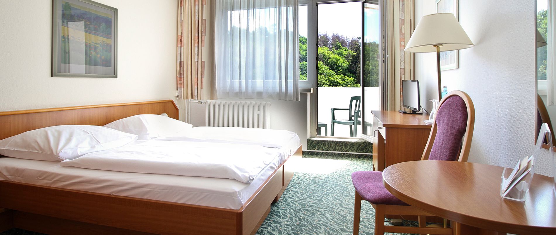 Helles Hotelzimmer mit Doppelbett, kleinem Tisch und Stuhl, Lampe und Zugang zu einem Balkon mit Blick ins Grüne.