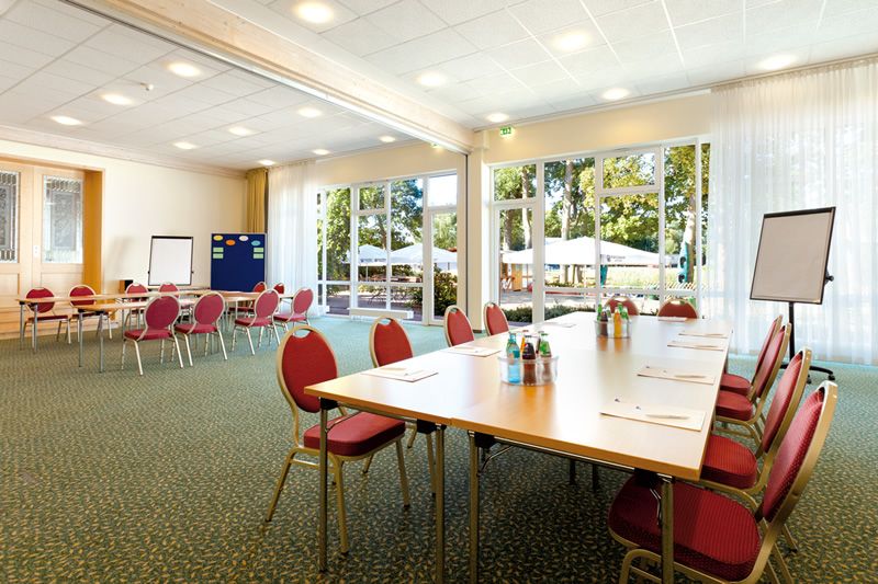 Konferenzraum mit Tischen und roten Stühlen, bereit für ein Meeting, mit Blick auf eine Terrasse durch große Fenster.