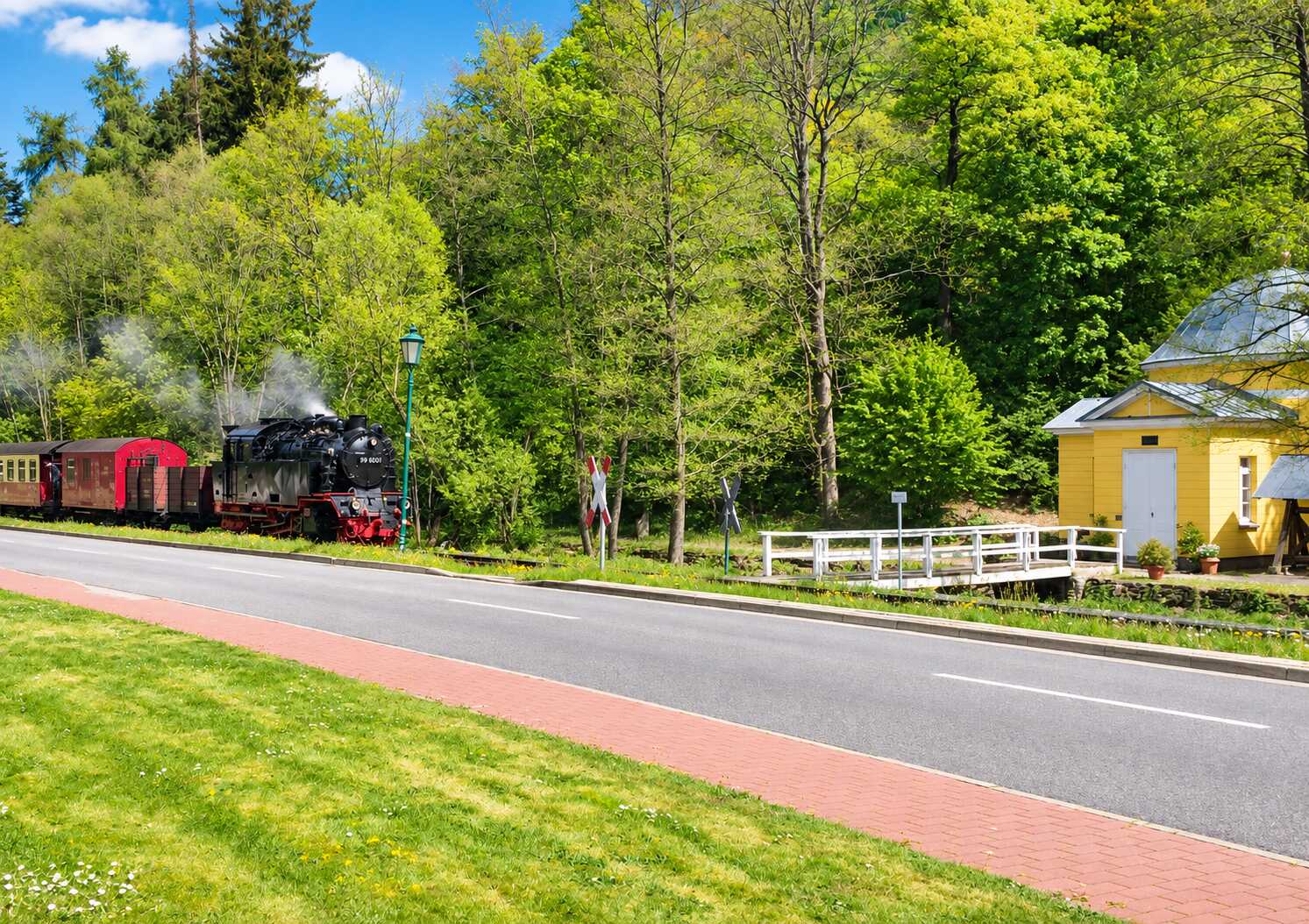 Eine Dampflokomotive mit angehängten Waggons fährt an einem gelben Gebäude vorbei, umgeben von Bäumen.