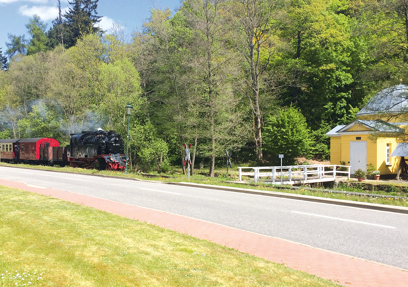 Eine Dampflokomotive mit angehängten Waggons fährt an einem gelben Gebäude vorbei, umgeben von Bäumen.
